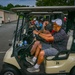 A line of Yamaha golf carts, numbered and staged in a parking area, are loaded with golfers preparing for a shotgun-start tournament or group outing. The foreground cart (#24) seats two men wearing caps and casual golf attire, with water bottles and scorecards visible. A person in a green shirt walks in the background near a storage building.