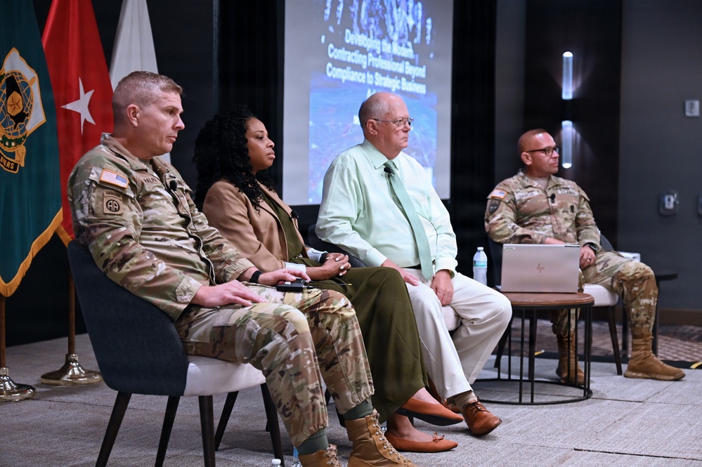 A panel discussion or briefing is taking place in a modern conference or auditorium setting, featuring four individuals seated in a row facing an audience. Three participants are wearing U.S. Army military uniforms in OCP (Operational Camouflage Pattern), while one civilian male is dressed in business casual attire. A presentation slide visible in the background reads 'Developing the Modern Contracting Professional Beyond Compliance to Strategic Business.'