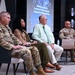 A panel discussion or briefing is taking place in a modern conference or auditorium setting, featuring four individuals seated in a row facing an audience. Three participants are wearing U.S. Army military uniforms in OCP (Operational Camouflage Pattern), while one civilian male is dressed in business casual attire. A presentation slide visible in the background reads 'Developing the Modern Contracting Professional Beyond Compliance to Strategic Business.'