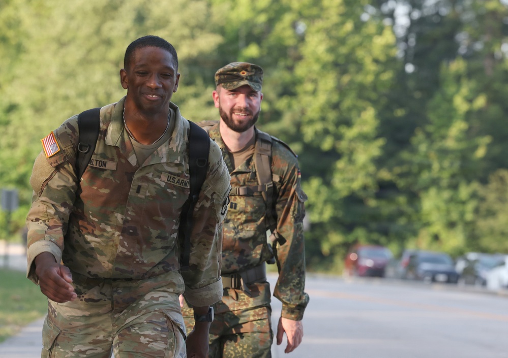 Two military personnel are walking along a road, both wearing different national military uniforms and carrying backpacks. The soldier in the foreground wears a U.S. Army OCP (Operational Camouflage Pattern) uniform with an American flag patch and a name tape reading 'LTON' (partially visible). The soldier in the background wears what appears to be a German Bundeswehr Flecktarn camouflage uniform with a matching field cap.