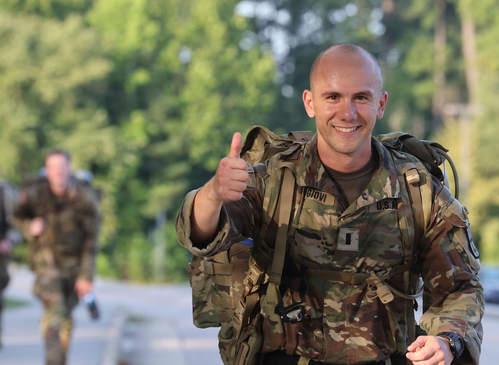 A U.S. Army soldier in OCP (Operational Camouflage Pattern) uniform is giving a thumbs-up gesture while carrying a rucksack, appearing to be mid-ruck march or physical training event. The soldier is smiling broadly and appears to be in good spirits despite the physical exertion. Additional soldiers in similar uniforms are visible in the blurred background.