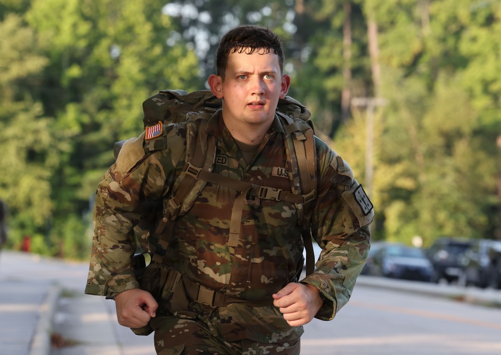 A U.S. Army soldier in OCP (Operational Camouflage Pattern) uniform is running while carrying a large military rucksack, likely during a ruck march or physical fitness event. The soldier appears visibly exerted, with sweat-dampened hair and a focused expression. The setting appears to be a paved road or path near a wooded area, consistent with a military installation.