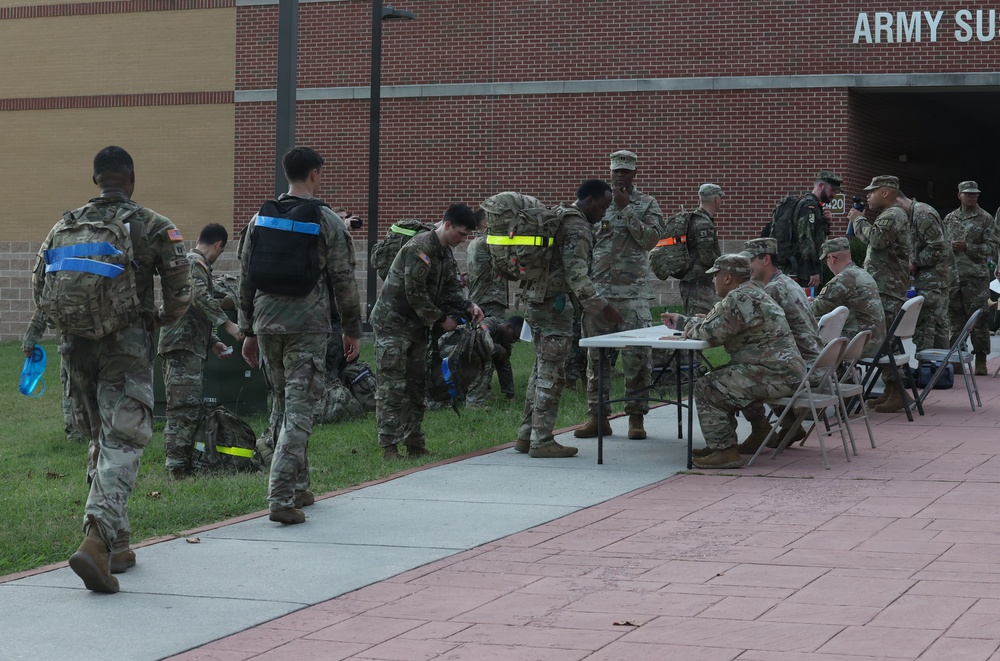 A group of U.S. Army soldiers in camouflage (OCP/MultiCam) uniforms are gathered outside a brick building partially labeled 'ARMY SU...' Several soldiers are carrying rucksacks marked with colored tape (blue, yellow, orange), suggesting a training exercise or accountability formation. A processing or check-in table staffed by seated soldiers is set up along a paved walkway.