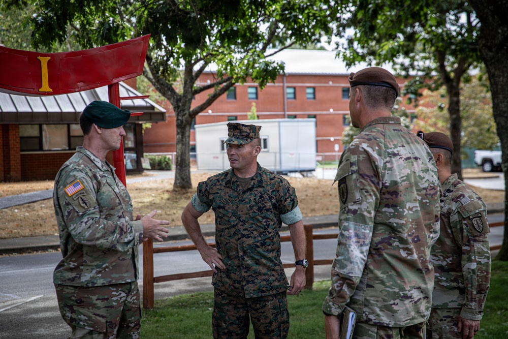 Four military personnel in different uniforms are engaged in a conversation outdoors on what appears to be a military installation. The group includes at least one U.S. Marine in MARPAT woodland camouflage and multiple U.S. Army soldiers in OCP (Operational Camouflage Pattern) uniforms with various beret colors. A red military unit insignia structure is visible in the background.