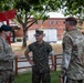 Four military personnel in different uniforms are engaged in a conversation outdoors on what appears to be a military installation. The group includes at least one U.S. Marine in MARPAT woodland camouflage and multiple U.S. Army soldiers in OCP (Operational Camouflage Pattern) uniforms with various beret colors. A red military unit insignia structure is visible in the background.