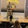 A U.S. Army soldier in OCP (Operational Camouflage Pattern) uniform is delivering a presentation or briefing on a stage at what appears to be the U.S. Army Quartermaster School. He stands at a wooden podium in front of a large display featuring the Quartermaster Corps insignia. Several soldiers seated in an auditorium-style setting are watching the presentation.