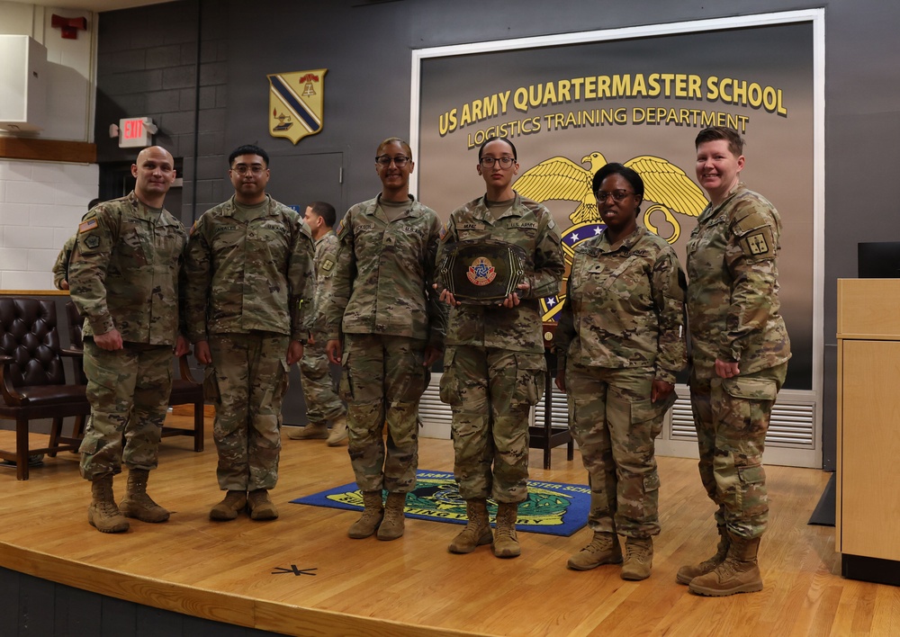 A group of seven U.S. Army soldiers in OCP (Operational Camouflage Pattern) uniforms pose for a formal photo on a stage at the US Army Quartermaster School, Logistics Training Department. One soldier in the center is holding what appears to be an award or trophy with a military insignia. The setting suggests an official recognition or graduation ceremony.