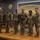 A group of seven U.S. Army soldiers in OCP (Operational Camouflage Pattern) uniforms pose for a formal photo on a stage at the US Army Quartermaster School, Logistics Training Department. One soldier in the center is holding what appears to be an award or trophy with a military insignia. The setting suggests an official recognition or graduation ceremony.