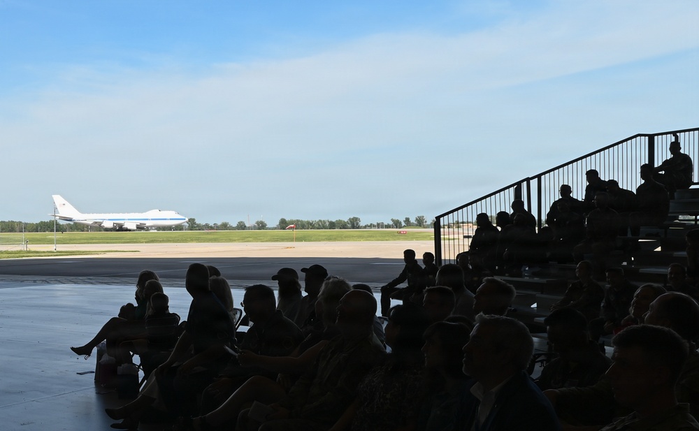 A crowd of people, silhouetted in the foreground, are seated in bleachers and on the ground inside or near a hangar, watching a large white aircraft on a runway or tarmac in the distance. The aircraft appears to be a wide-body jet, possibly a Boeing 747 variant, with a distinctive blue stripe livery. The setting appears to be a military or civilian airshow or aviation event on a clear, sunny day.