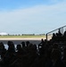 A crowd of people, silhouetted in the foreground, are seated in bleachers and on the ground inside or near a hangar, watching a large white aircraft on a runway or tarmac in the distance. The aircraft appears to be a wide-body jet, possibly a Boeing 747 variant, with a distinctive blue stripe livery. The setting appears to be a military or civilian airshow or aviation event on a clear, sunny day.