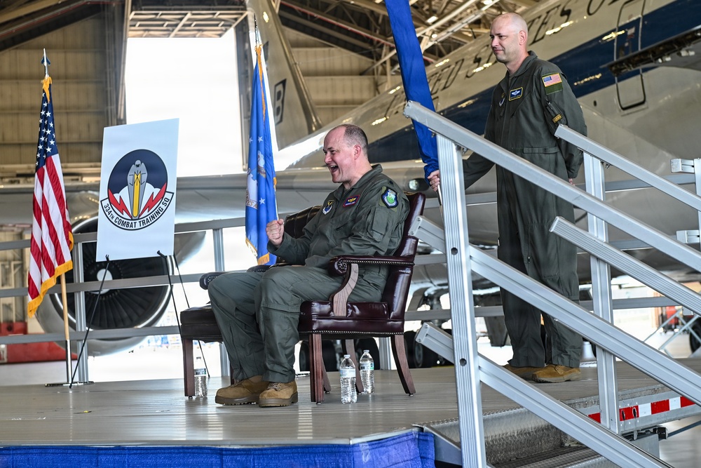 A military ceremony taking place inside an aircraft hangar, likely a change of command or activation ceremony for the 564th Combat Training Squadron, as indicated by the unit banner visible on stage. Two personnel in olive green flight suits are on an elevated stage platform, with one seated holding a blue guidon flag and another standing on adjacent stairs. An American flag and a light blue military flag are also present alongside a large military aircraft in the background.