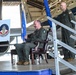 A military ceremony taking place inside an aircraft hangar, likely a change of command or activation ceremony for the 564th Combat Training Squadron, as indicated by the unit banner visible on stage. Two personnel in olive green flight suits are on an elevated stage platform, with one seated holding a blue guidon flag and another standing on adjacent stairs. An American flag and a light blue military flag are also present alongside a large military aircraft in the background.