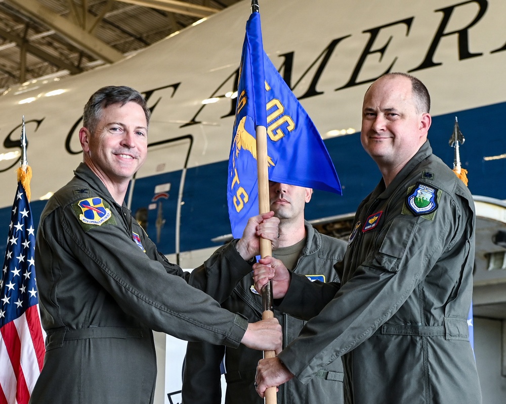 A military change of command ceremony is taking place inside an aircraft hangar, with two officers in olive green flight suits exchanging a blue unit guidon flag. A third service member holds the flag staff between them during the formal transfer. A large aircraft with partial text visible ('...AMER...') serves as the backdrop, along with an American flag.