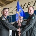 A military change of command ceremony is taking place inside an aircraft hangar, with two officers in olive green flight suits exchanging a blue unit guidon flag. A third service member holds the flag staff between them during the formal transfer. A large aircraft with partial text visible ('...AMER...') serves as the backdrop, along with an American flag.
