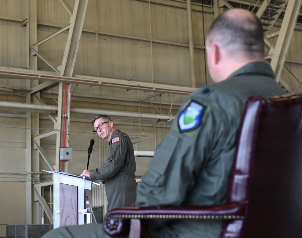 A military ceremony taking place inside an aircraft hangar, where one uniformed officer is speaking at a podium while another sits in a formal chair in the foreground. Both individuals are wearing olive green flight suits with military patches, suggesting a formal change of command or official military ceremony. The industrial hangar structure with steel beams and yellow framework is visible in the background.