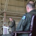 A military ceremony taking place inside an aircraft hangar, where one uniformed officer is speaking at a podium while another sits in a formal chair in the foreground. Both individuals are wearing olive green flight suits with military patches, suggesting a formal change of command or official military ceremony. The industrial hangar structure with steel beams and yellow framework is visible in the background.