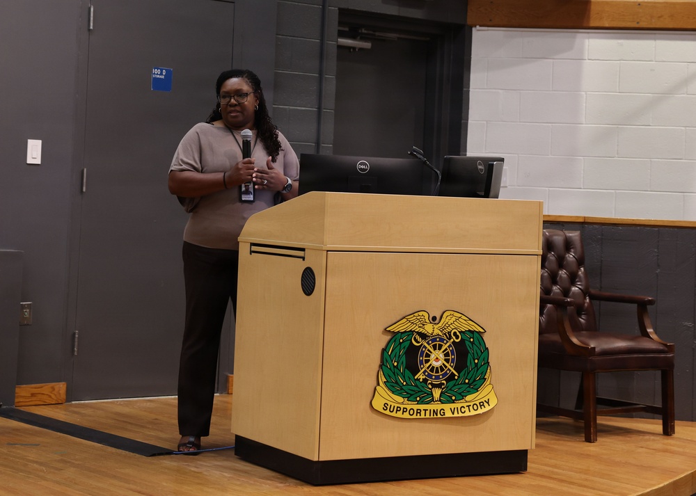 A woman stands at a wooden podium holding a microphone and appears to be giving a presentation or speech. The podium displays a military insignia with the motto 'Supporting Victory,' indicating a U.S. Army Sustainment Command or related military organization setting. A Dell laptop is visible on the podium, and the setting appears to be an auditorium or multipurpose room.