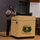 A woman stands at a wooden podium holding a microphone and appears to be giving a presentation or speech. The podium displays a military insignia with the motto 'Supporting Victory,' indicating a U.S. Army Sustainment Command or related military organization setting. A Dell laptop is visible on the podium, and the setting appears to be an auditorium or multipurpose room.