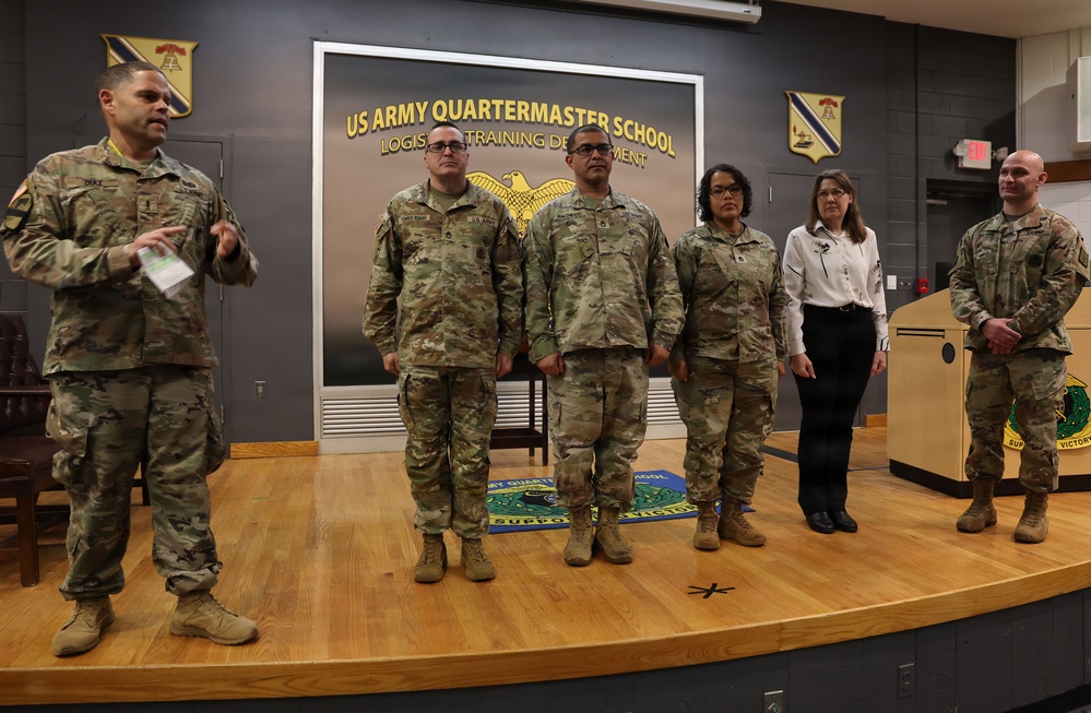 A group of six individuals stand on a stage at the US Army Quartermaster School, Logistics Training Department. Five are wearing US Army OCP (Operational Camouflage Pattern) uniforms, while one civilian woman in business attire stands among them. The setting appears to be a formal military ceremony or presentation event.