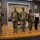 A group of six individuals stand on a stage at the US Army Quartermaster School, Logistics Training Department. Five are wearing US Army OCP (Operational Camouflage Pattern) uniforms, while one civilian woman in business attire stands among them. The setting appears to be a formal military ceremony or presentation event.