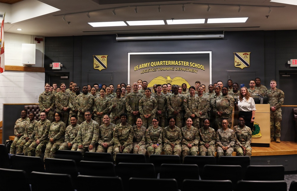 A large group of U.S. Army personnel pose for a formal group photograph inside an auditorium or briefing room at the US Army Quartermaster School, Logistics Training Department. The majority of individuals are wearing Army Combat Uniforms (ACU/OCP pattern), with one civilian woman visible near a podium on the right. The setting features institutional signage, unit crests, and a staged seating arrangement typical of a military school environment.