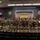 A large group of U.S. Army personnel pose for a formal group photograph inside an auditorium or briefing room at the US Army Quartermaster School, Logistics Training Department. The majority of individuals are wearing Army Combat Uniforms (ACU/OCP pattern), with one civilian woman visible near a podium on the right. The setting features institutional signage, unit crests, and a staged seating arrangement typical of a military school environment.