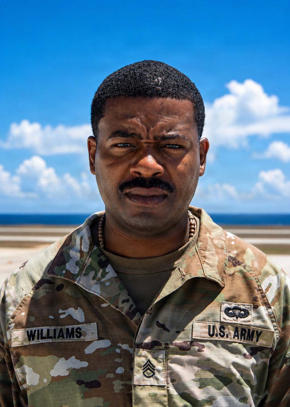 A U.S. Army Staff Sergeant named Williams poses for a portrait photograph outdoors. He is wearing Operational Camouflage Pattern (OCP) uniform with visible name tape, branch tape, rank insignia, and a parachutist badge. The background shows a blue sky with clouds and what appears to be a coastal or waterfront environment.