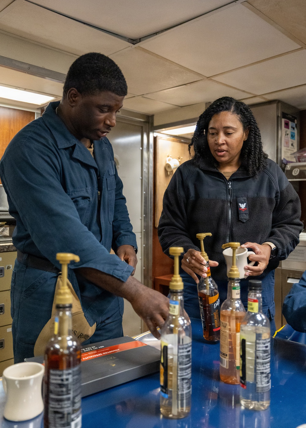 Two individuals in what appears to be a military or naval vessel workspace are working with multiple bottles equipped with pump dispensers on a blue-surfaced counter. The person on the left is wearing navy blue coveralls, while the person on the right is wearing a black fleece jacket with a visible insignia patch. They appear to be organizing or preparing syrup/flavoring bottles, possibly for a coffee or beverage station.