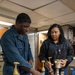 Two individuals in what appears to be a military or naval vessel workspace are working with multiple bottles equipped with pump dispensers on a blue-surfaced counter. The person on the left is wearing navy blue coveralls, while the person on the right is wearing a black fleece jacket with a visible insignia patch. They appear to be organizing or preparing syrup/flavoring bottles, possibly for a coffee or beverage station.