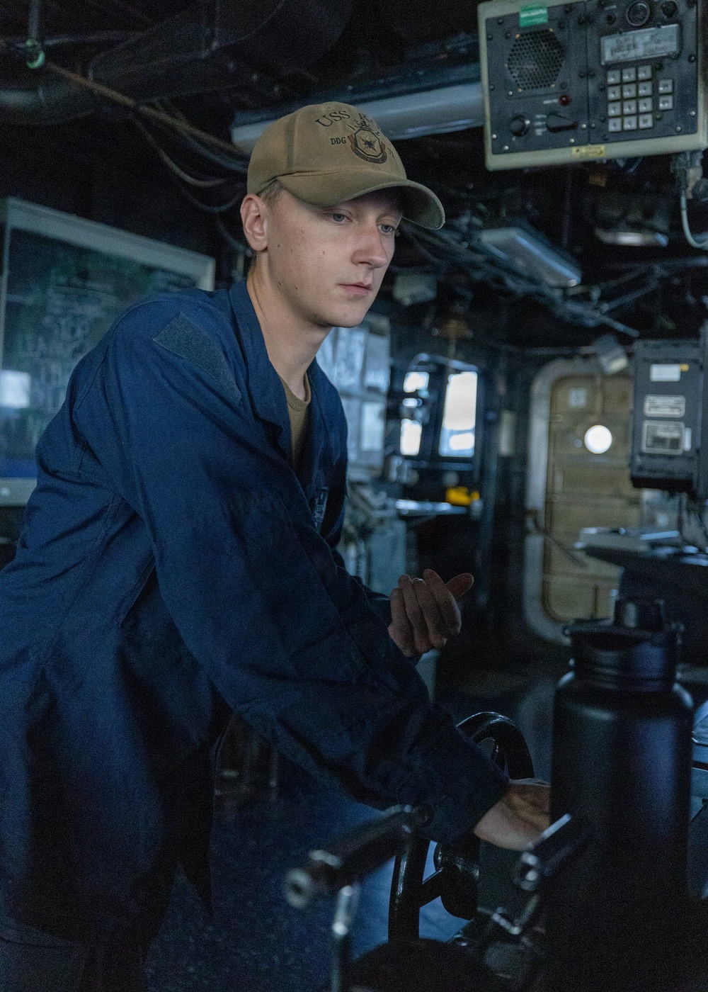 A young male sailor in blue Navy coveralls (utilities) stands at what appears to be a ship's helm or navigation station aboard a U.S. Navy vessel. He is wearing a tan/olive baseball cap with 'USS' and 'DDG' visible, indicating assignment to a destroyer-class ship. The setting is the bridge or pilothouse of a naval vessel, with various electronic equipment and instrumentation visible in the background.