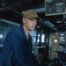 A young male sailor in blue Navy coveralls (utilities) stands at what appears to be a ship's helm or navigation station aboard a U.S. Navy vessel. He is wearing a tan/olive baseball cap with 'USS' and 'DDG' visible, indicating assignment to a destroyer-class ship. The setting is the bridge or pilothouse of a naval vessel, with various electronic equipment and instrumentation visible in the background.