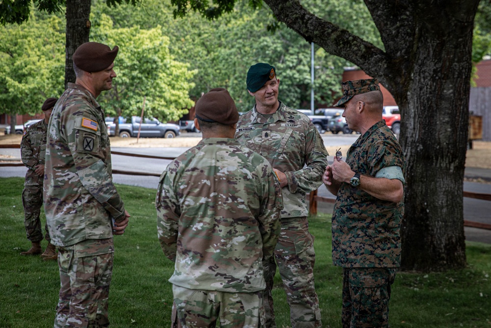 A group of U.S. military personnel from multiple branches are engaged in an informal outdoor conversation on a grassy area near trees. The group includes U.S. Army soldiers wearing OCP uniforms with green and brown berets, and at least one U.S. Marine in MARPAT digital camouflage. The setting appears to be a military installation, with vehicles and buildings visible in the background.