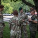 A group of U.S. military personnel from multiple branches are engaged in an informal outdoor conversation on a grassy area near trees. The group includes U.S. Army soldiers wearing OCP uniforms with green and brown berets, and at least one U.S. Marine in MARPAT digital camouflage. The setting appears to be a military installation, with vehicles and buildings visible in the background.