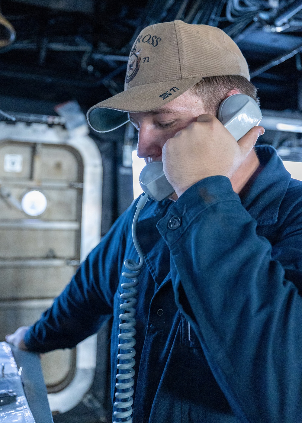 A U.S. Navy sailor is using a corded shipboard telephone while standing at a console aboard a naval vessel. The sailor is wearing a tan ball cap with 'ROSS' and 'DDG 71' markings, identifying the ship as USS Ross (DDG-71), an Arleigh Burke-class guided-missile destroyer. The setting appears to be an interior compartment or bridge area of the ship.