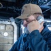 A U.S. Navy sailor is using a corded shipboard telephone while standing at a console aboard a naval vessel. The sailor is wearing a tan ball cap with 'ROSS' and 'DDG 71' markings, identifying the ship as USS Ross (DDG-71), an Arleigh Burke-class guided-missile destroyer. The setting appears to be an interior compartment or bridge area of the ship.