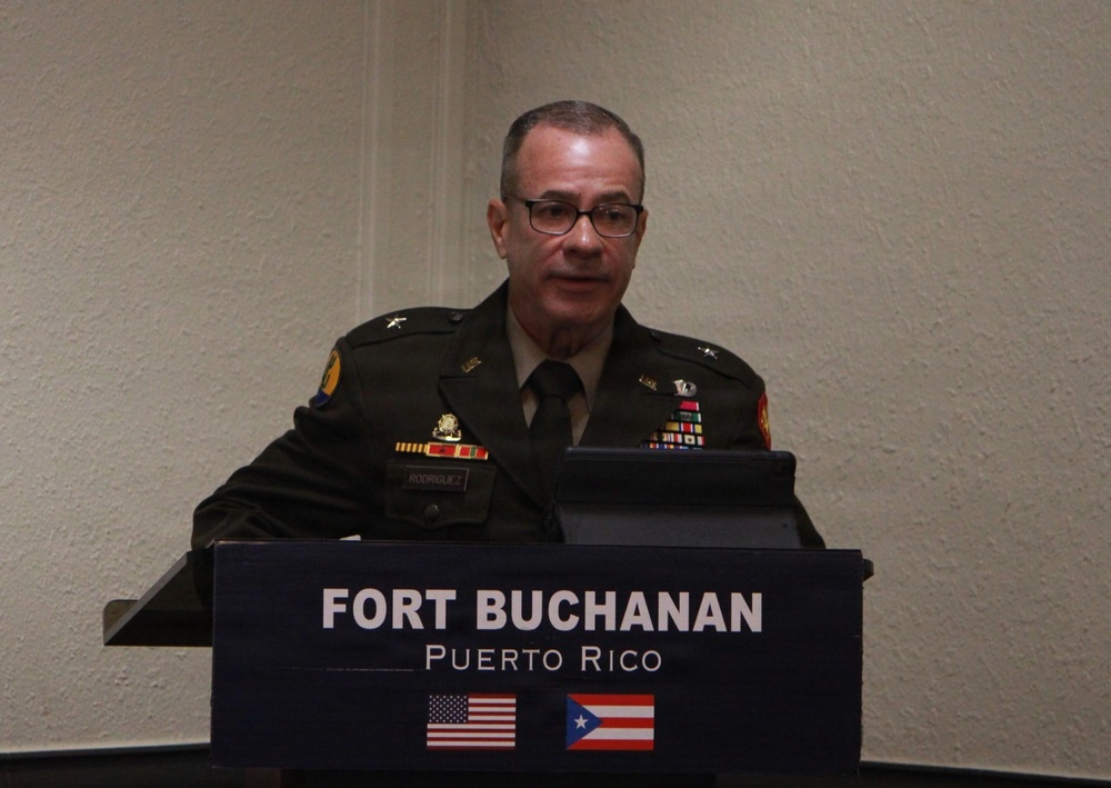 A U.S. Army officer in dress uniform stands at a podium bearing the 'Fort Buchanan, Puerto Rico' insignia with both the American and Puerto Rican flags displayed. The officer appears to be delivering a speech or briefing at an official military event. Multiple service ribbons and medals are visible on the uniform, indicating significant military service.