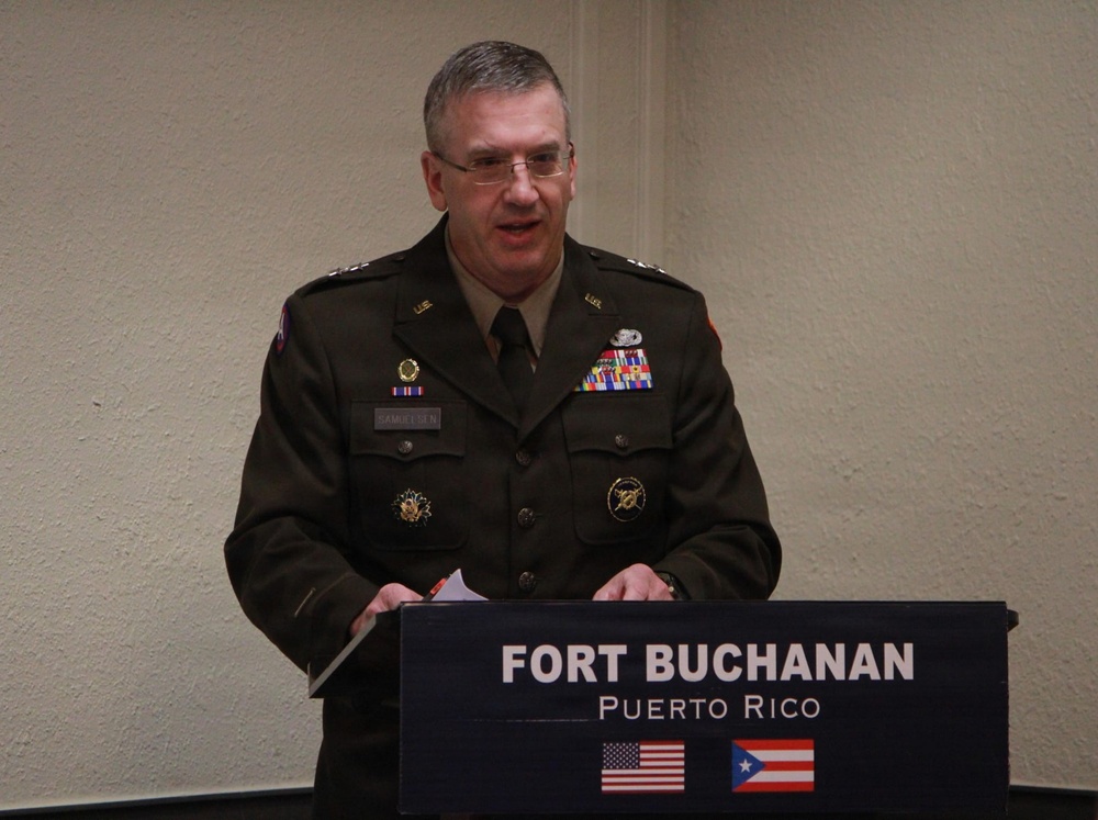 A U.S. Army officer identified by nameplate as 'Samuelsen' stands at a podium bearing the 'Fort Buchanan, Puerto Rico' insignia. He is wearing a Class A dress uniform with multiple ribbons, badges, and rank insignia, and appears to be delivering a speech or address. The podium displays both the U.S. flag and the Puerto Rico flag.