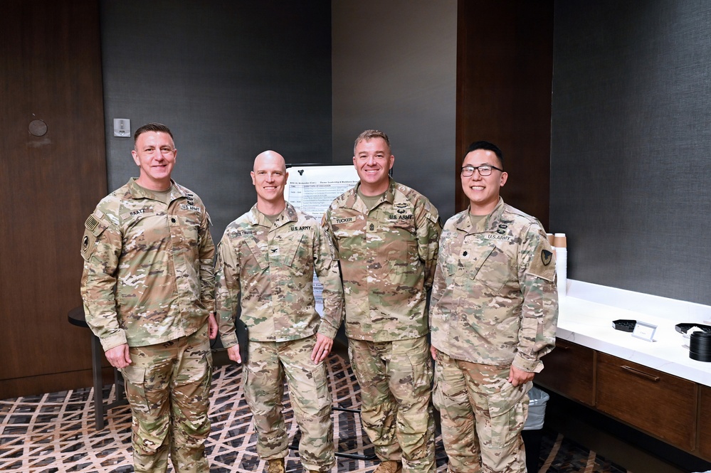 Four U.S. Army soldiers in OCP (Operational Camouflage Pattern) uniforms are posing together for a group photo in what appears to be a conference or meeting room. Name tapes are visible on their uniforms, with readable names including 'PAATZ', 'STRUSS' (or similar), 'TUCKER', and a fourth soldier on the right. The setting suggests a formal military event or training conference.