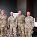 Four U.S. Army soldiers in OCP (Operational Camouflage Pattern) uniforms are posing together for a group photo in what appears to be a conference or meeting room. Name tapes are visible on their uniforms, with readable names including 'PAATZ', 'STRUSS' (or similar), 'TUCKER', and a fourth soldier on the right. The setting suggests a formal military event or training conference.