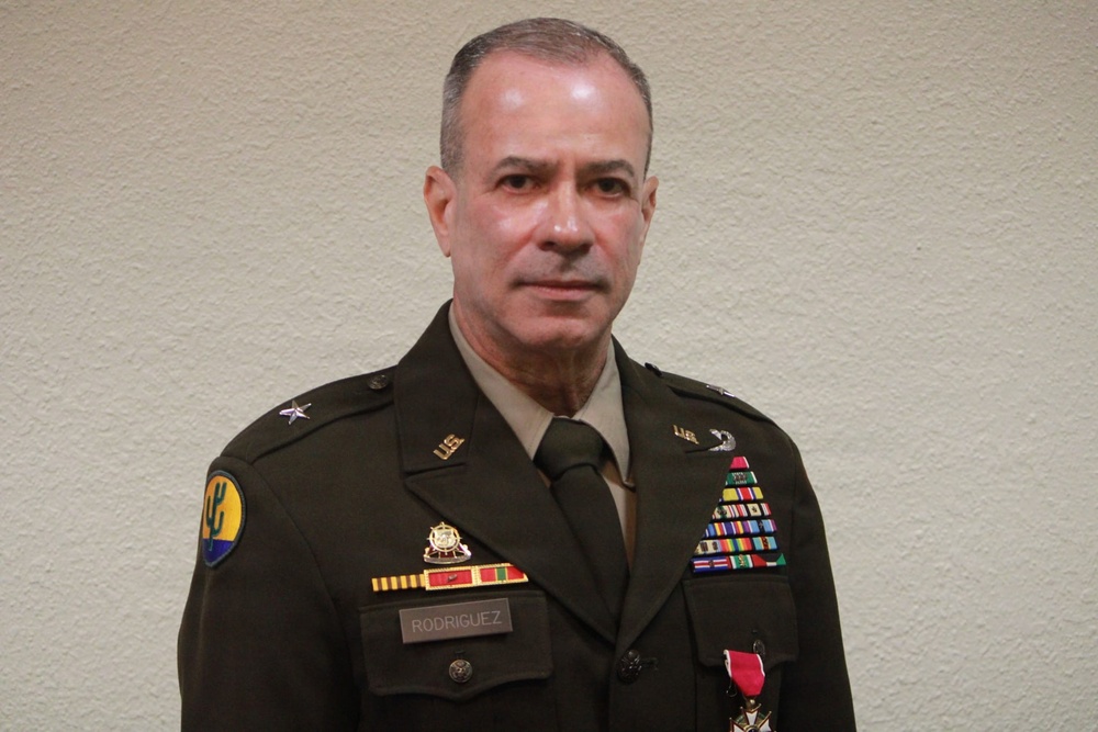 A U.S. Army officer in dress green uniform is photographed against a plain beige wall in what appears to be an official or semi-formal portrait setting. The name tag reads 'RODRIGUEZ' and the uniform displays numerous medals, ribbons, and insignia indicating significant military service. The subject appears to be a senior officer based on rank insignia and decorations visible.