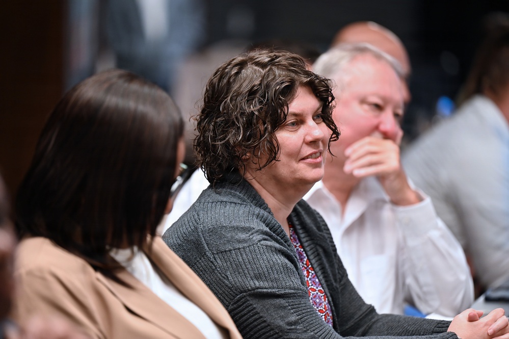 The image shows a group of people seated together, likely at a conference, meeting, or formal event. The central figure is a person with dark curly hair wearing a gray cardigan over a floral top, appearing engaged and slightly smiling. The background contains additional attendees in soft focus, suggesting a professional or academic gathering.
