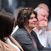 The image shows a group of people seated together, likely at a conference, meeting, or formal event. The central figure is a person with dark curly hair wearing a gray cardigan over a floral top, appearing engaged and slightly smiling. The background contains additional attendees in soft focus, suggesting a professional or academic gathering.