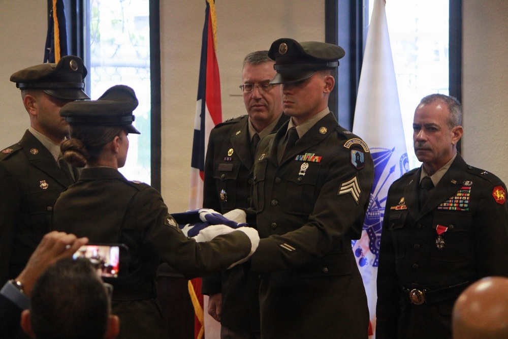 A formal U.S. Army military ceremony is taking place indoors, featuring a flag folding or presentation ritual. Several soldiers in Army Service Uniforms (Class A/green) are participating, with one soldier holding a folded American flag while another receives it. Audience members are visible in the foreground, with one person recording the event on a smartphone.