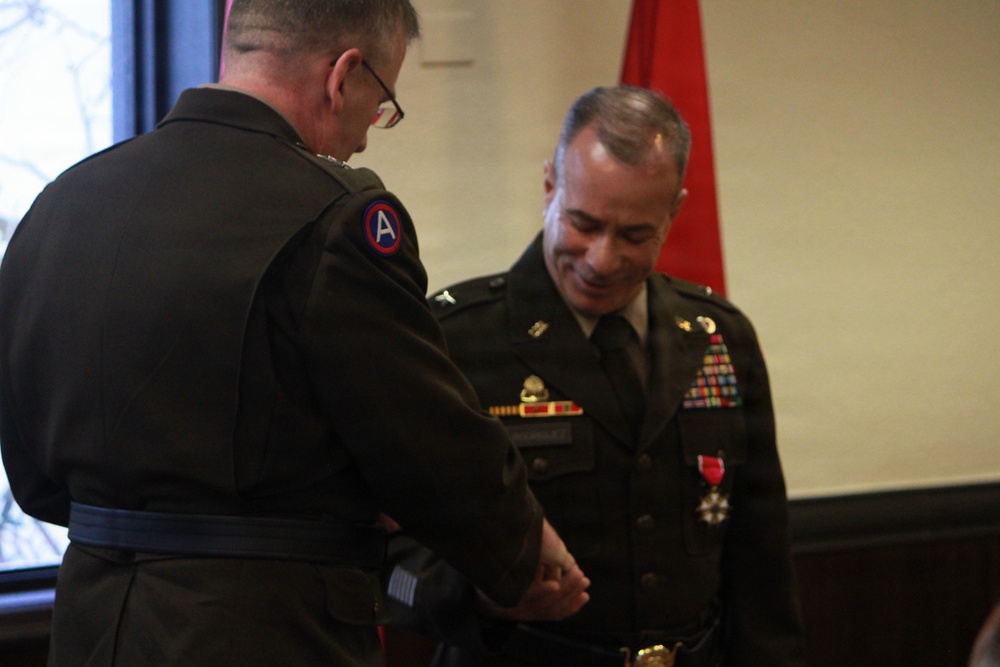 Two U.S. Army officers in Class A green dress uniforms are engaged in what appears to be a military awards or decoration ceremony. The officer on the right is already wearing a medal with a red ribbon, while the officer on the left appears to be pinning or adjusting something on the right officer's uniform. A red military flag or guidon is visible in the background.
