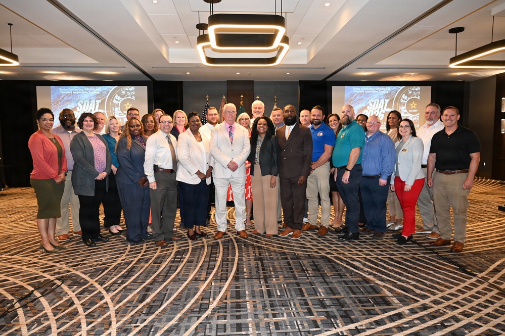 A large group of approximately 30 professionals poses for a formal group photograph in what appears to be a hotel conference or ballroom setting. The event appears to be associated with 'SDAT' (visible on projection screens in the background), which appears to be a U.S. Army-related program based on the Army logo visible on the screens. The group is diverse in gender and ethnicity, dressed in business and business-casual attire.
