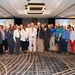 A large group of approximately 30 professionals poses for a formal group photograph in what appears to be a hotel conference or ballroom setting. The event appears to be associated with 'SDAT' (visible on projection screens in the background), which appears to be a U.S. Army-related program based on the Army logo visible on the screens. The group is diverse in gender and ethnicity, dressed in business and business-casual attire.