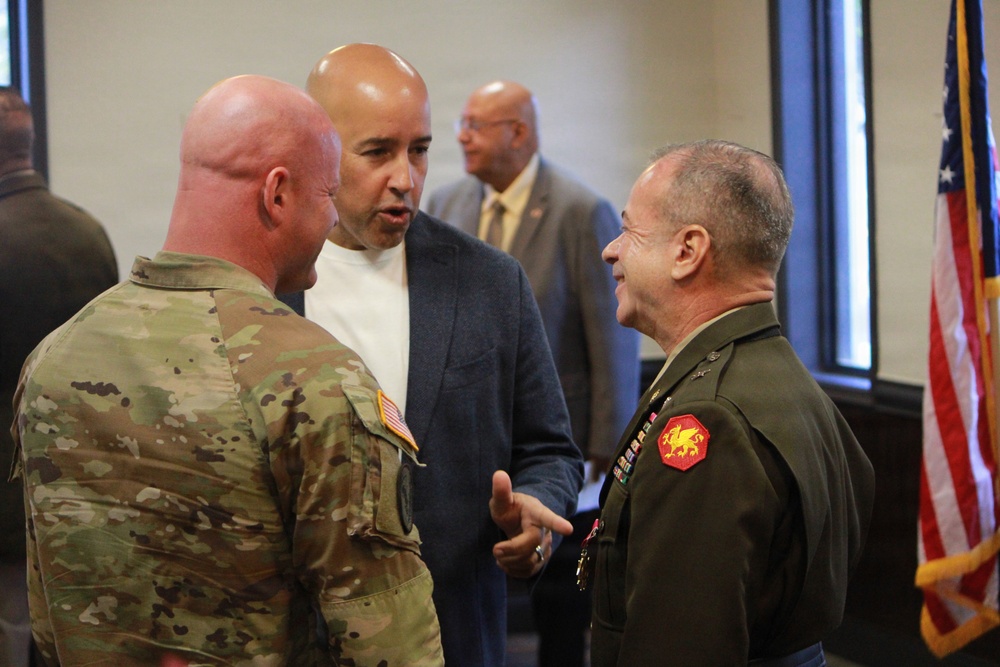 Three men are engaged in conversation in what appears to be an official or ceremonial indoor setting. Two are in military uniforms — one in Army OCP (Operational Camouflage Pattern) and one in Marine Corps dress greens with medals and a unit patch — while the third is in civilian business casual attire. A U.S. flag is visible in the background along with additional individuals.