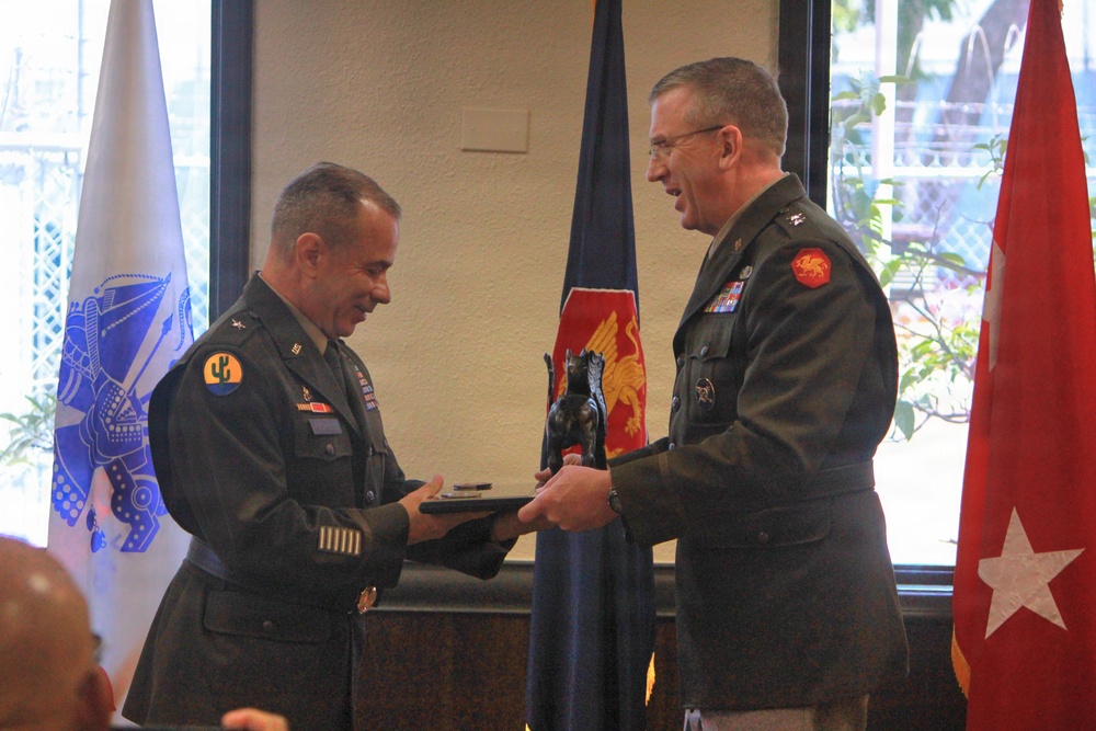 Two U.S. Army officers in Army Service Uniforms (ASUs) are exchanging what appears to be a plaque or award during a formal military ceremony. Both wear unit patches and rank insignia; the officer on the left displays a cactus patch (likely 40th Infantry Division or similar) and the officer on the right wears a red dragon patch. Several military flags and a general officer flag (red with white star) are visible in the background.