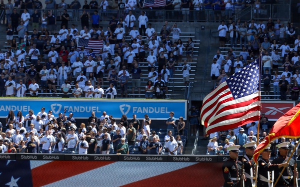 A military color guard ceremony is taking place on a baseball field, likely during a pre-game presentation at what appears to be Yankee Stadium. Several uniformed military personnel are holding the American flag and a red military branch flag on the outfield grass. The stadium is filled with a large crowd of spectators, many dressed in white and navy team colors.