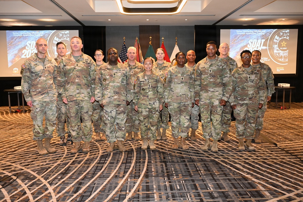 A group of approximately 14 U.S. Army personnel in OCP (Operational Camouflage Pattern) uniforms pose for a formal group photograph in what appears to be a hotel conference room or ballroom. The event is identified by projected screens as 'SDAT 2024' - Senior Contracting Officials and Directors Acquisition Training. Multiple U.S. and Army flags are displayed prominently in the background.